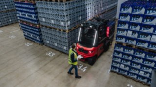 Egy láthatósági mellényt viselő személy egy olyan állványfolyosó mellett sétál el, ahonnan épp egy targonca tolat ki.