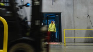 Egy védőruhát viselő férfi kilép egy ajtón egy üzemcsarnokban. Egy emelővillás targonca előre halad. Egy védőruhát viselő férfi kilép egy ajtón egy üzemcsarnokban. Egy emelővillás targonca előre halad.