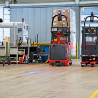 Két L-MATIC típusú automata elektromos raklapemelő árukat szállít a csomagolósor és az átfolyó állványos raktár között.