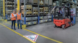 Linde TruckSpot jelet vetít a raktárban Két ember megy a raktárban, útjukat oldalról keresztezi egy targonca