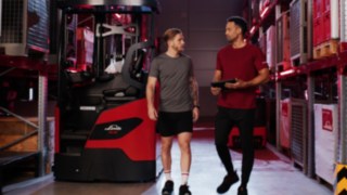 Két sportruhás személy áll a Linde Anyagmozgatás Ri16 sorozatú tolóoszlopos targoncája előtt, és egyikük táblagépet tart a kezében.