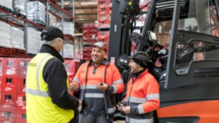A Linde targoncáinak kényelme és biztonsága növeli a járművezetők munkával való elégedettségét és motiváltságát. Három ember beszélget egy targonca előtt.